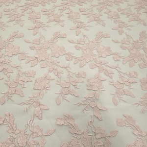 Puede incluir: Primer plano de una tela de encaje floral rosa claro. La tela presenta un intrincado patrón de flores y hojas bordadas, creando una superficie delicada y texturizada. El diseño está distribuido uniformemente por toda la tela.
