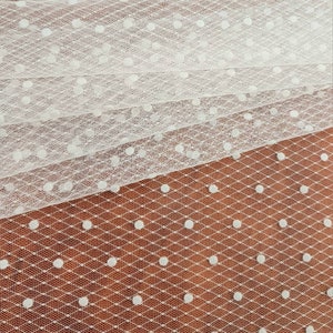 May include: White tulle fabric with small white polka dots. The fabric is folded in layers, showing the pattern.