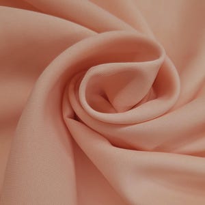 Puede incluir: Primer plano de una tela color melocotón con pliegues suaves y una textura sutil. La tela parece lisa y se drapea suavemente, creando una sensación de fluidez y movimiento. El color es un tono cálido y apagado.