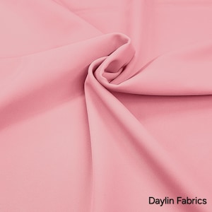 Pode incluir: Um close-up de um tecido rosa macio com dobras e uma textura suave. O tecido é de cor sólida e parece ser um material leve. O texto "Daylin Fabrics" está no canto inferior direito.