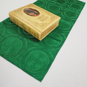 May include: A green fabric book cover with a repeating pattern of circles and crosses. The cover is folded in half and a Holy Bible book is placed on top of it.