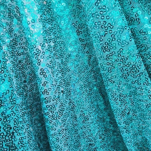 Op de afbeelding: Close-up van turquoise stof met een paillettenpatroon. De stof is gevouwen en gedrapeerd, waardoor de textuur van de pailletten zichtbaar wordt.