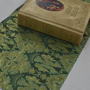 May include: A vintage Holy Bible sits on a dark green and gold patterned runner. The Bible has a gold cover with embossed text and an oval illustration. The runner features a repeating design of angels and ornate floral elements.