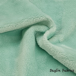 May include: A close-up of a soft, light green fabric with a fuzzy texture. The fabric is folded and draped, showing its texture and drape. The text "Daylin Fabrics" is visible in the bottom right corner.