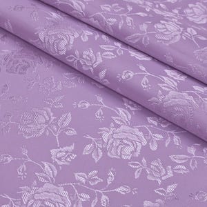 Könnte beinhalten: Nahaufnahme eines lavendelfarbenen Stoffes mit einem floralen Rosenmuster. Der Stoff ist gefaltet und zeigt die Textur und das Design. Die Rosen und Blätter sind in einem helleren Lavendelton gehalten, was einen subtilen Kontrast erzeugt.