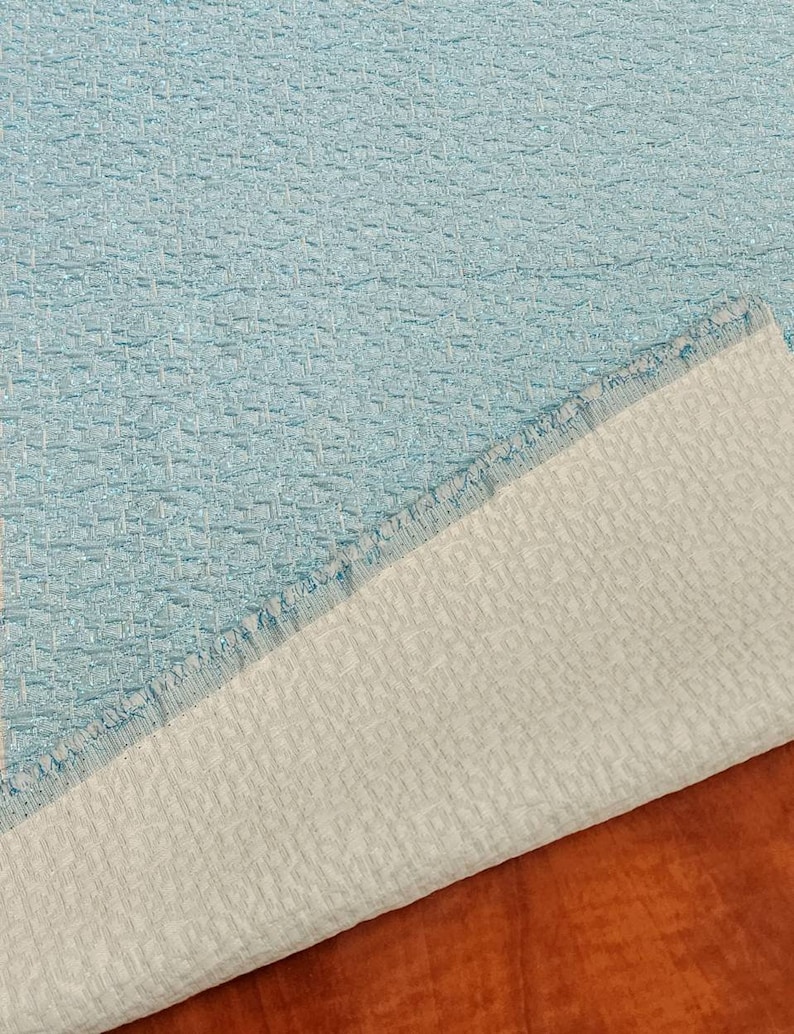 May include: Close-up of a light blue fabric with a textured woven pattern. The fabric is folded over, revealing a white backing.