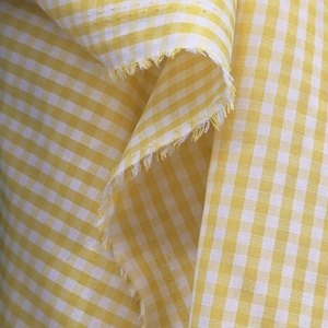 Pode incluir: Um pedaço de tecido com um padrão clássico de xadrez vichy em amarelo e branco. O material está dobrado para revelar uma borda crua e desfiada com fios soltos visíveis ao longo da costura, mostrando a textura tecida do tecido xadrez.