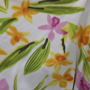 Op de afbeelding: Witte stof met een bloemenpatroon. Het patroon bevat oranje, roze en gele bloemen, en groene bladeren. Het ontwerp is een herhalend patroon van gestileerde bloemen en bladeren.