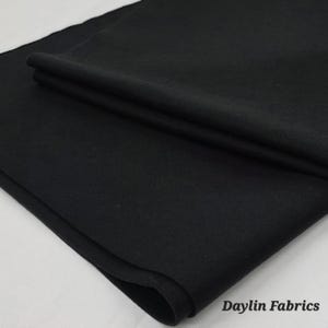 May include: A folded piece of black fabric, possibly felt or a similar material, with a soft, matte appearance. The fabric is layered, showing the texture and depth of the material. The words "Daylin Fabrics" are in the bottom right corner.
