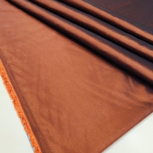 May include: A close-up shot of a folded, rust-colored fabric with a subtle sheen. The fabric has a soft texture and is gathered in several folds, creating shadows and highlights. The edge of the fabric has a frayed, orange trim.