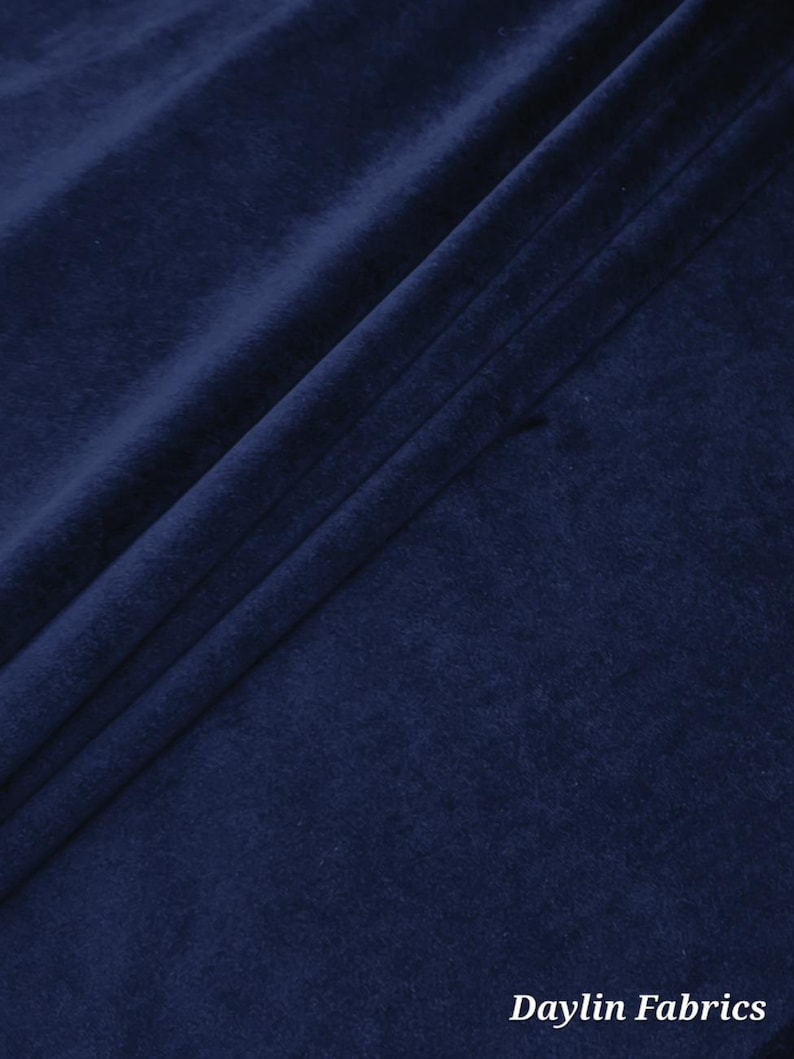 May include: Close-up of a deep navy blue fabric with a soft, velvety texture. The fabric is draped, creating folds and shadows that highlight its plush appearance. The text "Daylin Fabrics" is visible in the bottom right corner.