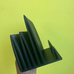 May include: A black, angular desk organizer with multiple vertical slots. The organizer is made of a solid, matte material and is held against a yellow background. The design features a series of angled dividers, creating individual compartments for storage.