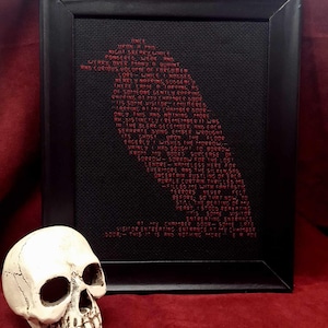 May include: A black framed embroidery with red thread depicting a poem about a visitor entering a chamber. The poem reads: "DE SORTEONE GENTLY SOPPING POPPING 01 MEMBER NOOR IS SOME VISITORY MILTERED SUNG ONLY TUTS AND NOTHING MORE RM DISTINE FLY REMEMBER IT WAS IN THE BLERK DECEMBER AND EACH SEPARATE DYING EMBER WROUGHT OPON THE FLOOR ERGERLY I WISHED THE MORROW URINLY I HAD SOUGHT TO BORROW FROM MY BOOKS BURGERSE OF SORROW- SORROW FOR THE LOS LENDRE KAMELESS HERE FOR EVERMORE AND THE SILKEN SRE UNCERTAIN BUSTLING BEERCA PURPLE CURTAIN THRILLED ME -FILLED ME WITH FANTASTIC TERRORS NEVER FELT BEFORE, 50 TURT NOW, TO STILL THE BERTING OF MY HEART STOOD REPEATING TIS SOME VISITOR ENTRERTING ENTRANCE 1 [MBER DOOR SOME URTE VISITOR ENTRERTING ENTRANCE ET MY Снимаея DOOR THIS IS RNA NOTHING MORE ER"