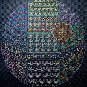 Puede incluir: Un diseño de bordado circular con fondo negro y patrones florales coloridos. El texto "Build Gardens, not Empires" está bordado en blanco alrededor del borde del círculo. El texto "Mimas Terraformed" está bordado en blanco en la parte inferior del círculo.