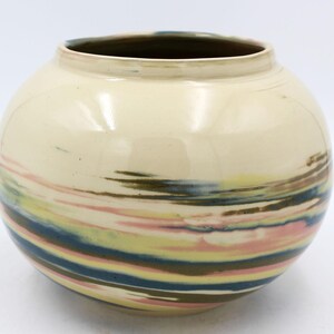 May include: A round ceramic vase with a cream-colored exterior and a dark interior. The vase features horizontal bands of color, including pink, blue, yellow, and brown, creating a swirled effect. The vase has a wide opening.