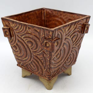 May include: A square, brown ceramic planter with a textured, swirling pattern. The planter has a tapered shape and sits on three small, tan feet. Decorative elements are present on the corners.