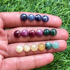 May include: A hand holds a collection of faceted gemstones in diverse colours. The gemstones include shades of blue, red, yellow, and green. The gems are round and have a polished, reflective surface, ideal for jewellery making.