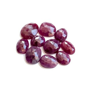 Puede incluir: Un grupo de diez piedras preciosas de rubí facetadas de diferentes tamaños y formas. Las piedras preciosas son de color rojo púrpura intenso y tienen un acabado pulido y brillante.
