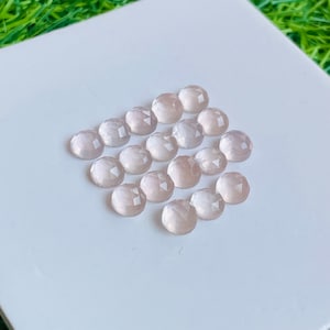 Könnte beinhalten: Eine Sammlung von 18 facettierten Cabochons aus hellrosa Edelsteinen. Die Cabochons sind in einem Halbkreis auf einer weißen Oberfläche angeordnet.