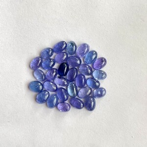 Op de afbeelding: Een verzameling ovale, gefacetteerde edelstenen in tinten blauw en paars. De stenen zijn in een losse cirkel op een witte achtergrond gerangschikt.