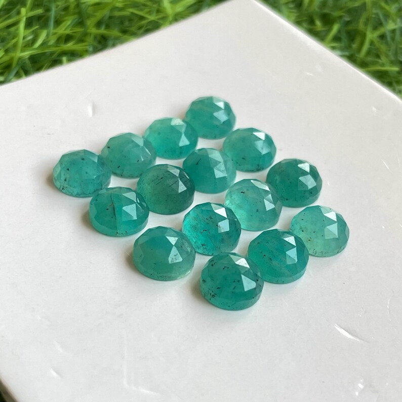 May include: A collection of 15 teal green faceted gemstone cabochons. The cabochons are round and have a smooth, polished surface.