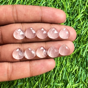 Könnte beinhalten: Eine Sammlung von acht rosa Rosenquarz-Facettensteinen in Tropfenform. Die Steine sind in zwei Reihen zu je vier auf einer Hand angeordnet.