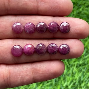 May include: Eight faceted ruby gemstones, each with a pink-red hue, are arranged in two rows of four on a hand. The gemstones are round and have a smooth, polished finish.