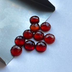 May include: A group of 11 red-brown gemstone cabochons, each with a faceted, rounded shape. The stones are arranged in a loose triangular pattern on a silver surface.