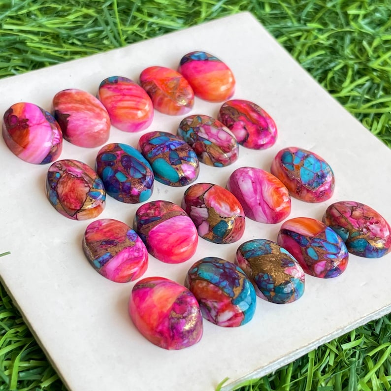 May include: A collection of oval-shaped gemstones in various colors, including pink, orange, blue, and gold. The gemstones are arranged on a white surface, with a green background. The stones have a marbled appearance.
