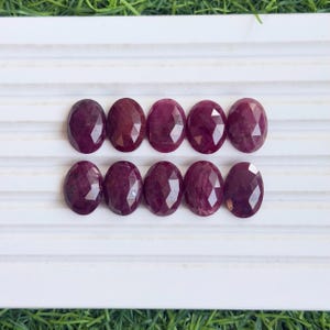 Puede incluir: Diez rubíes ovalados y facetados dispuestos en dos filas de cinco. Cada piedra preciosa muestra un tono magenta rojizo profundo y saturado con una superficie pulida de múltiples facetas que refleja la luz sobre sus bordes redondeados.