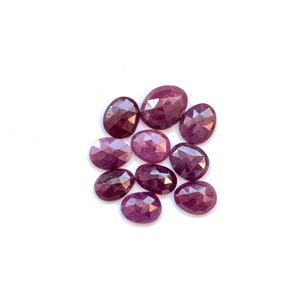 Puede incluir: Un grupo de diez piedras preciosas de rubí ovaladas facetadas en varios tonos de rosa y morado. Las piedras preciosas están dispuestas en un racimo sobre un fondo blanco.