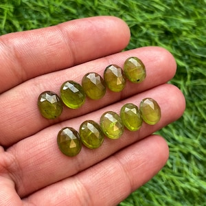 May include: A collection of nine faceted, oval-shaped peridot gemstones. The gems are a vibrant olive green color and are displayed against a background of green grass. Each stone has multiple facets that catch the light.