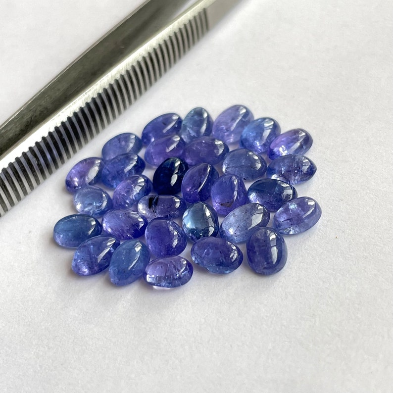 Op de afbeelding: Een groep ovale, blauwe edelstenen, waarschijnlijk tanzaniet, verspreid over een wit oppervlak. De edelstenen hebben verschillende tinten blauw, van licht tot donker.
