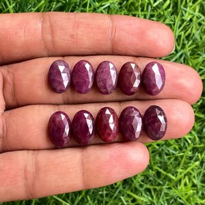 May include: Ten oval-shaped, faceted ruby gemstones. The rubies are a deep, rich red color, with a polished, reflective surface. The gemstones are arranged in two rows, displayed against a green background.