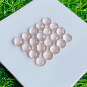 Könnte beinhalten: Eine Ansammlung von einundzwanzig hellrosa, facettierten Edelsteinen, die in einer rautenförmigen Anordnung angeordnet sind. Die runden Edelsteine haben eine mehrfach facettierte Oberfläche, die das Licht reflektiert. Die Edelsteine werden auf einer weißen quadratischen Oberfläche präsentiert.