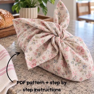 Puede incluir: Una envoltura de tela beige con un pequeño estampado floral rosa y azul, atada con un nudo en la parte superior. La imagen incluye una flecha negra que apunta a la tela con el texto: PDF pattern + step by step instructions.
