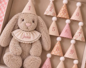 Puede incluir: Un conejo de peluche beige con orejas largas lleva un cuello color crema bordado con el nombre Janie y un gorro de fiesta con el número 1. Junto al conejo hay once gorros de fiesta adicionales numerados del 2 al 12, cada uno con un pompón blanco.
