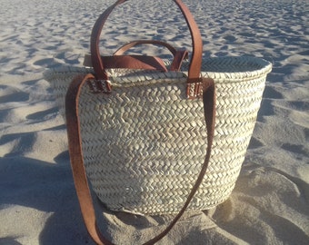 May include: Woven straw tote bag with brown leather handles and a shoulder strap. The bag has a rounded shape and is sitting on a sandy surface. The leather straps are attached to the bag with white stitching.