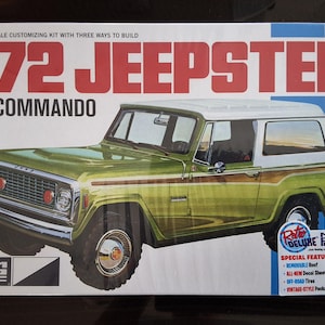 May include: A 1/25 scale model kit box for a '72 Jeepster Commando. The box features a green and white vehicle illustration with wood grain side panels. Text includes '72 JEEPSTER COMMANDO' and 'SPECIAL FEATURES' listing a removable roof.