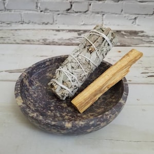 May include: A purple speckled stone bowl holds a white sage bundle tied with string and a piece of palo santo wood. The bowl sits on a white wooden surface with a white brick wall in the background.