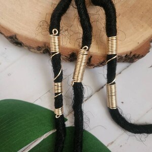 May include: Close-up of three black dreadlocks adorned with gold-colored wire wraps. The wraps are arranged in a spiral and cylindrical pattern, adding a decorative element to the hair. The background features a wooden surface and a green leaf.