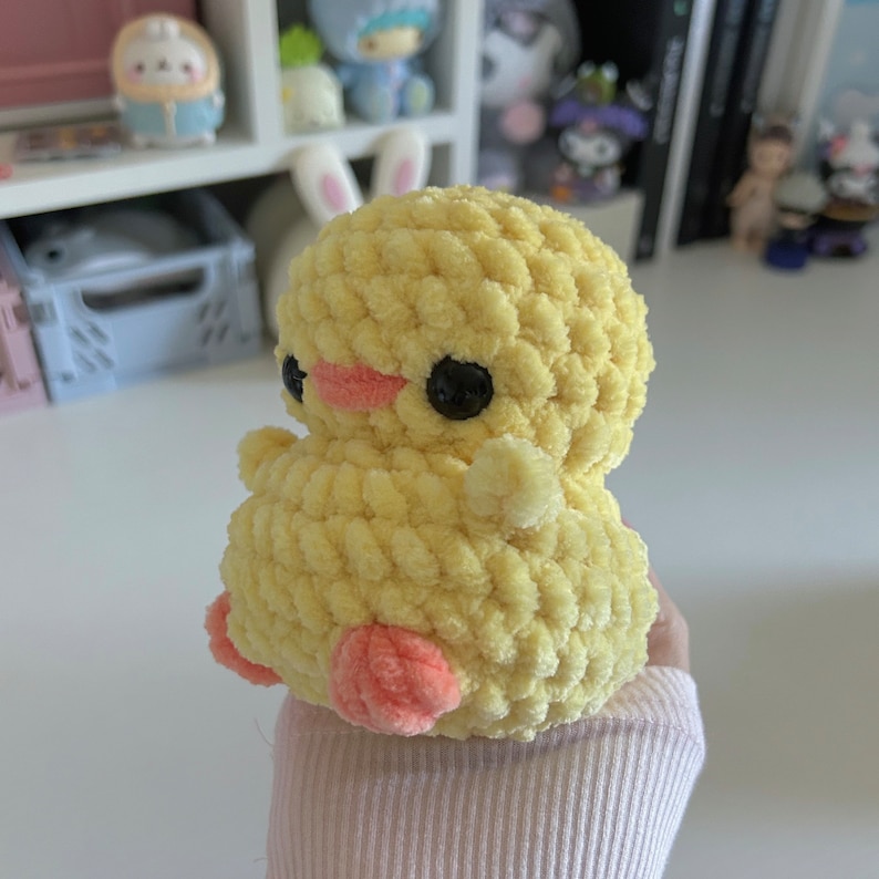 May include: A handmade, yellow crochet duckling with black eyes and a pink beak and feet. The plush toy is soft and fluffy, with a rounded body and small wings. This adorable duckling is a charming addition to any collection.