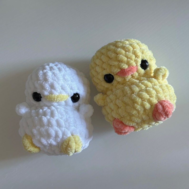 May include: Two handmade crochet ducklings. One is white with yellow beak and feet, the other is yellow with pink accents. Both have large black eyes, giving them a cute, cartoonish appearance. These soft toys are perfect for baby showers or nursery decor.