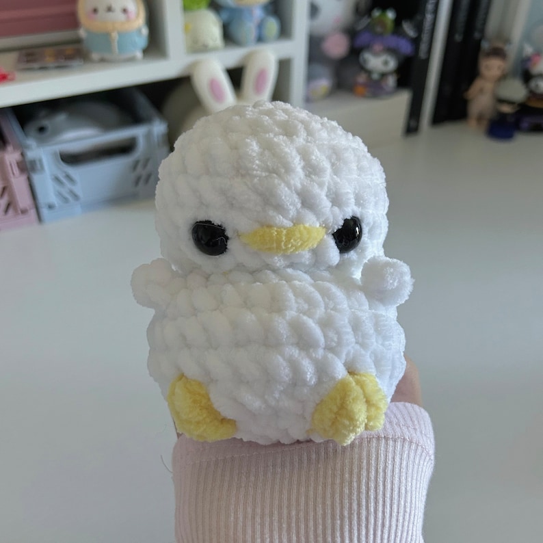 May include: A handmade white crochet duckling with black eyes and a yellow beak and feet. The plush toy has small wings and is made of soft, textured yarn. The duckling is a cute and cuddly item.