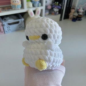 May include: A white crochet duck toy with black eyes and a yellow beak and feet. The soft toy is handmade and suitable for children or as a decorative item. The duck is held in a person's hand.