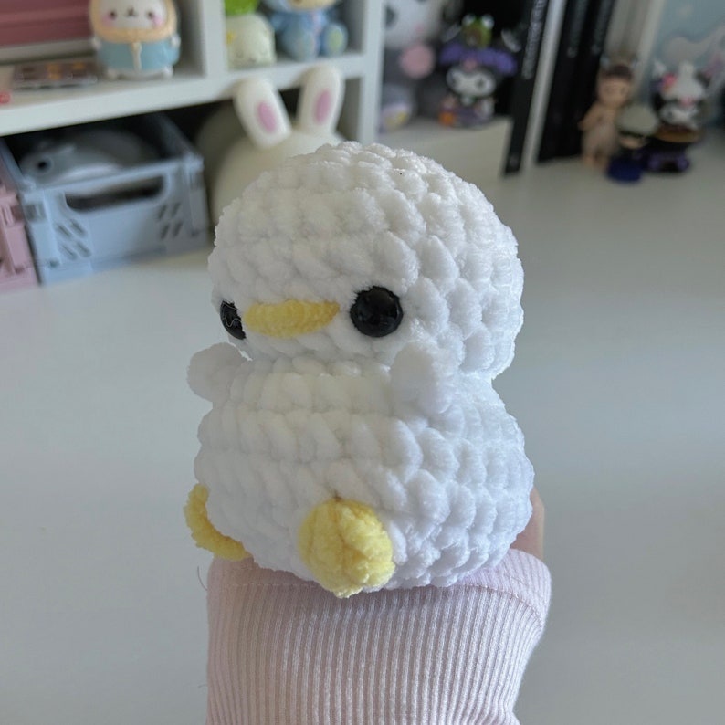 May include: A white crochet duck toy with black eyes and a yellow beak and feet. The soft toy is handmade and suitable for children or as a decorative item. The duck is held in a person's hand.