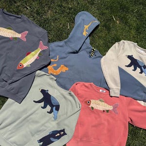 K&ouml;nnte beinhalten: Eine Auswahl an Sweatshirts und einem Hoodie in verschiedenen Farben, darunter Blau, Gr&uuml;n und Koralle. Die Sweatshirts zeigen applizierte Fisch- und B&auml;renmotive. Der Hoodie hat ein appliziertes Eidechsen-Design. Alle Artikel sind aus weichem Stoff.