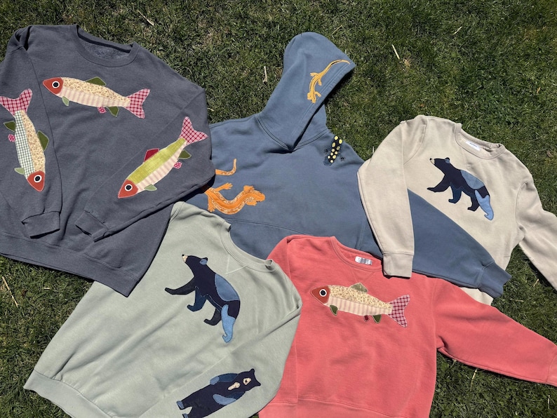 K&ouml;nnte beinhalten: Eine Auswahl an Sweatshirts und einem Hoodie in verschiedenen Farben, darunter Blau, Gr&uuml;n und Koralle. Die Sweatshirts zeigen applizierte Fisch- und B&auml;renmotive. Der Hoodie hat ein appliziertes Eidechsen-Design. Alle Artikel sind aus weichem Stoff.