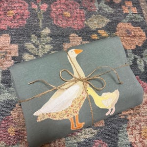 K&ouml;nnte beinhalten: Ein salbeigr&uuml;nes Stoffst&uuml;ck, das mit Bindfaden verschn&uuml;rt ist und eine applizierte Gans und ein G&auml;nsek&uuml;ken zeigt. Die Gans hat einen floral gemusterten K&ouml;rper, und das G&auml;nsek&uuml;ken ist gelb. Der Hintergrund ist ein floral gemusterter Teppich.