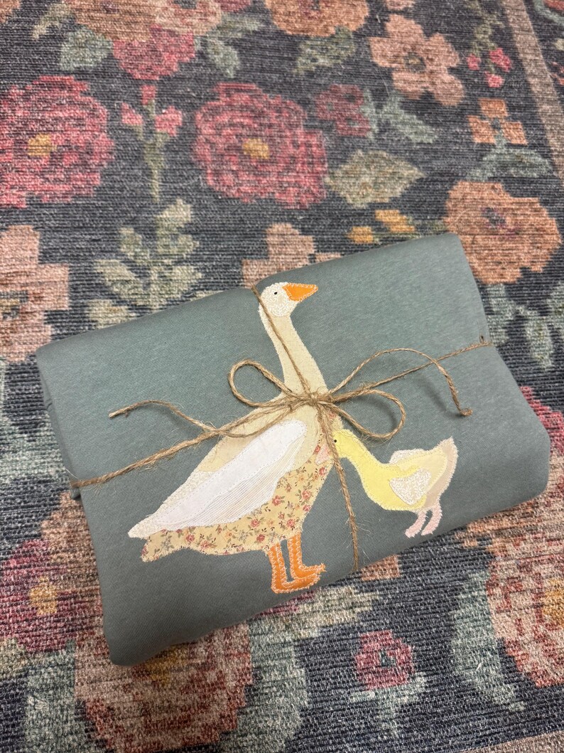K&ouml;nnte beinhalten: Ein salbeigr&uuml;nes Stoffst&uuml;ck, das mit Bindfaden verschn&uuml;rt ist und eine applizierte Gans und ein G&auml;nsek&uuml;ken zeigt. Die Gans hat einen floral gemusterten K&ouml;rper, und das G&auml;nsek&uuml;ken ist gelb. Der Hintergrund ist ein floral gemusterter Teppich.